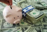 Piggy bank being filled with a coin next to stacks of U.S. dollar bills