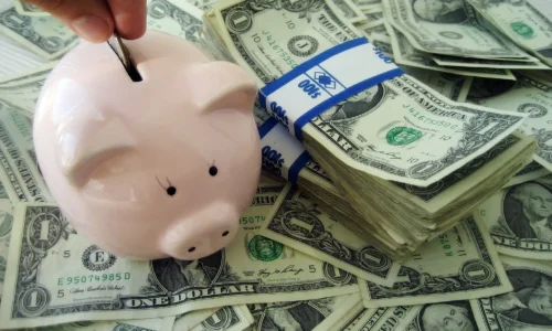 Piggy bank being filled with a coin next to stacks of U.S. dollar bills