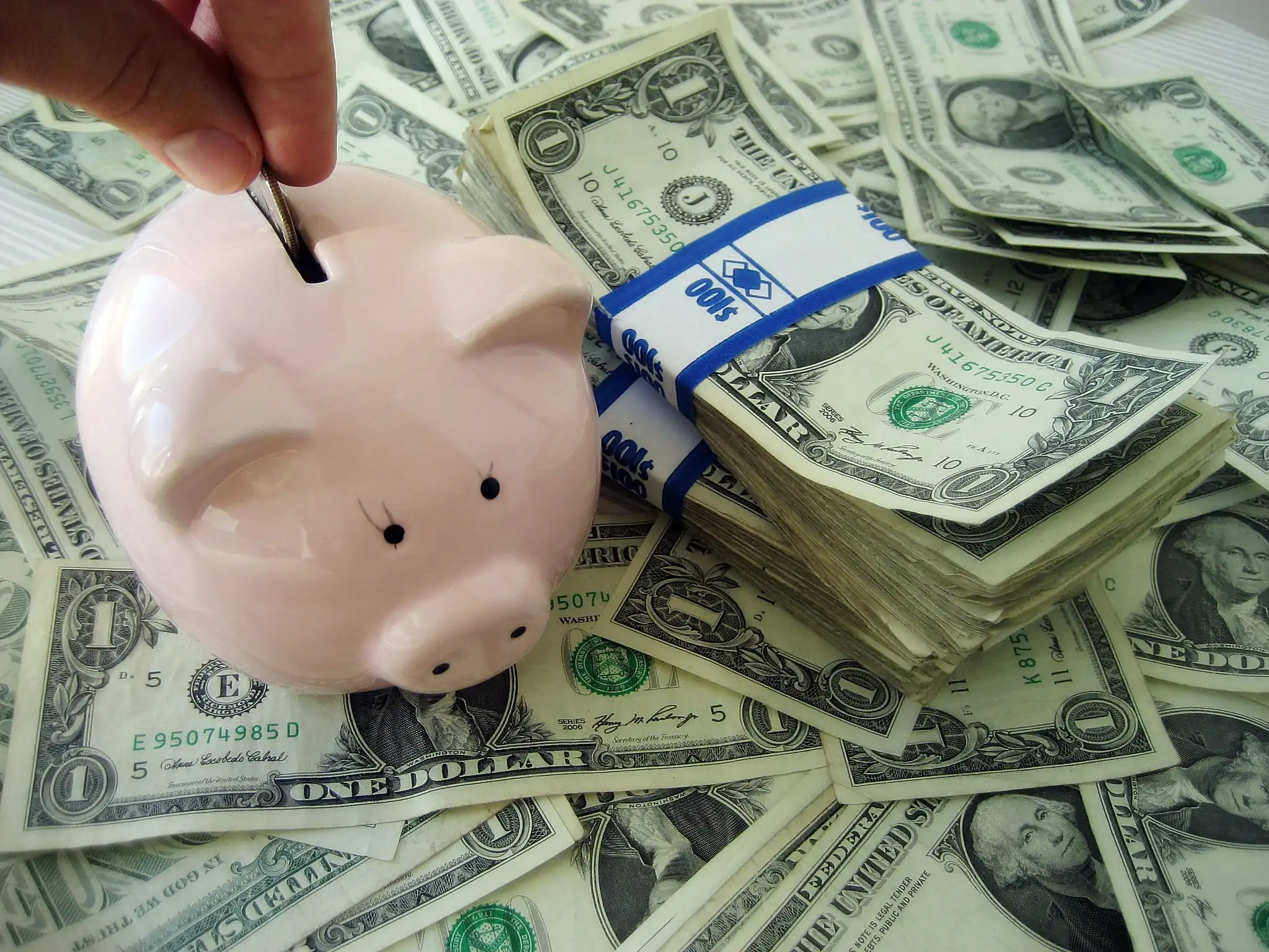 Piggy bank being filled with a coin next to stacks of U.S. dollar bills