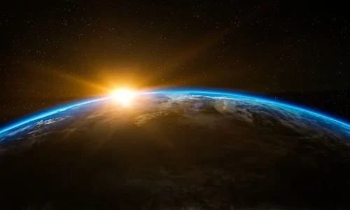 A view of a planet's curved surface with sunlight emerging over the horizon and stars in the background
