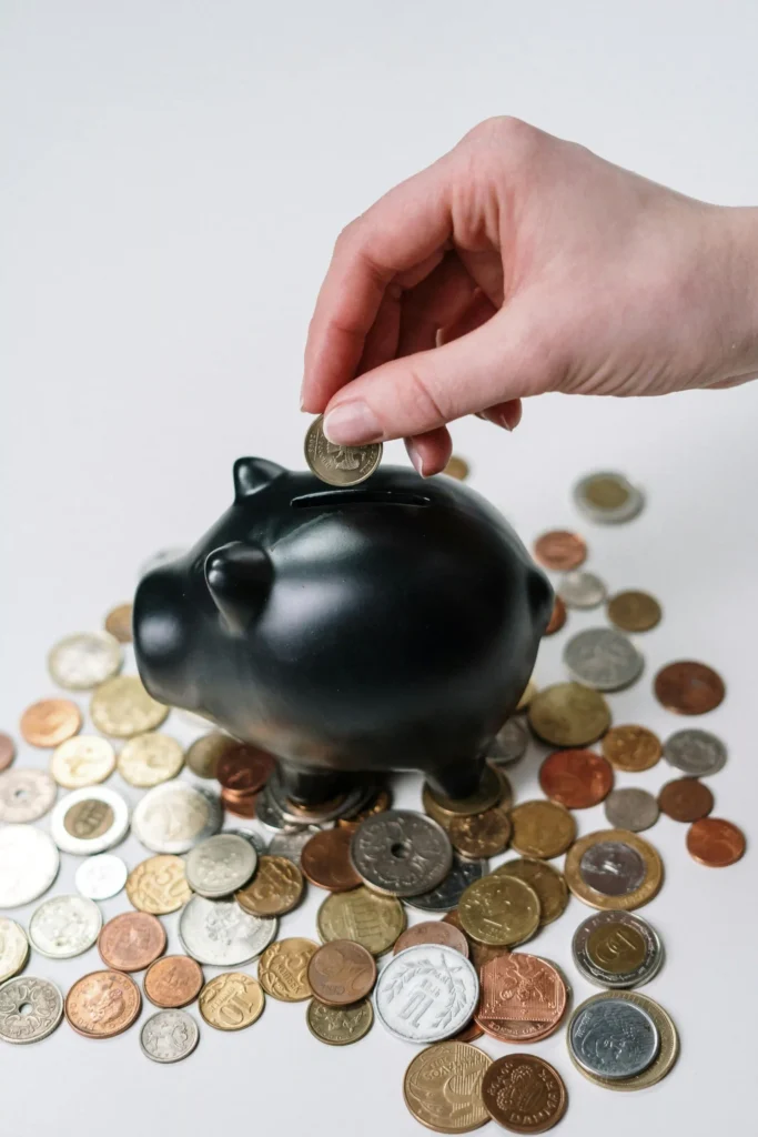 Hand placing a coin into a black piggy bank surrounded by various coins on a white background